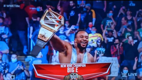 Big E holds up the WWE championship belt as fans stand and applaud him.