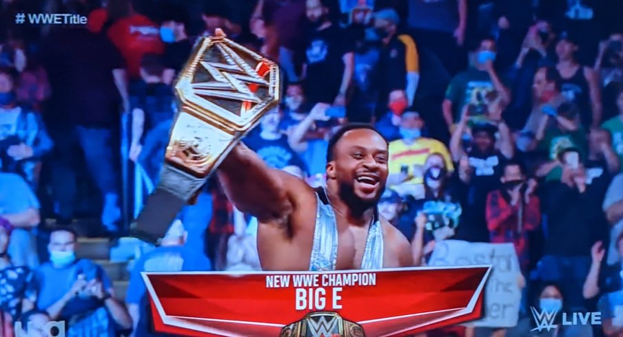 Big E holds up the WWE championship belt as fans stand and applaud him.